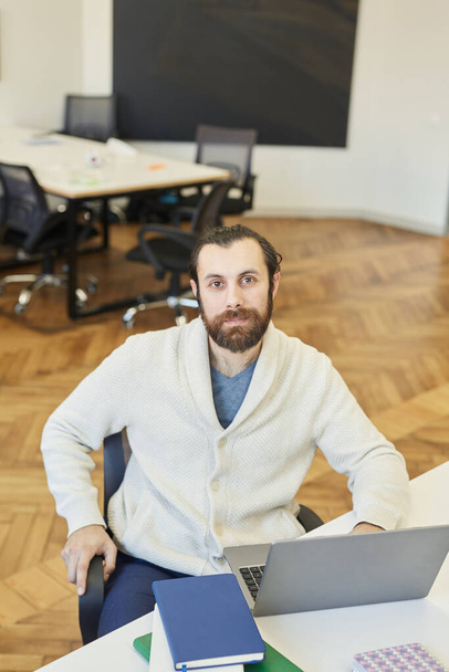 Portrait vertical à grand angle du jeune homme caucasien moderne avec barbe sur le visage portant une tenue confortable assis au bureau, regardant la caméra - Photo, image