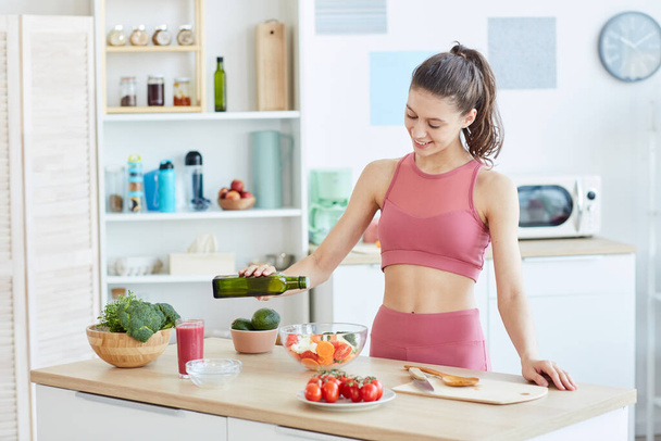 Derékig érő portré fitt fiatal nő öntés olívaolaj saláta főzés közben fitness ételek konyha belsejében, másolás hely - Fotó, kép