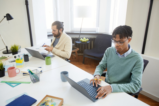Οριζόντια λήψη δύο σύγχρονων υπαλλήλων γραφείου που κάθονται στα τραπέζια και κάνουν τη δουλειά τους χρησιμοποιώντας φορητούς υπολογιστές - Φωτογραφία, εικόνα