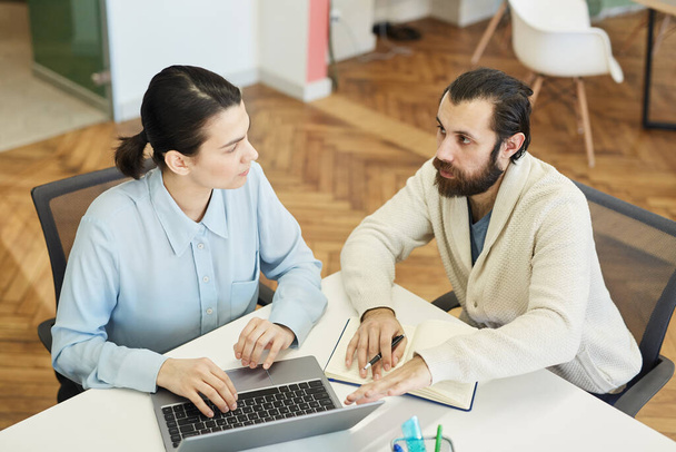 Young Caucasian man with beard on face and attractive young woman sitting together at office desk working on project, high angle view shot - Foto, Bild