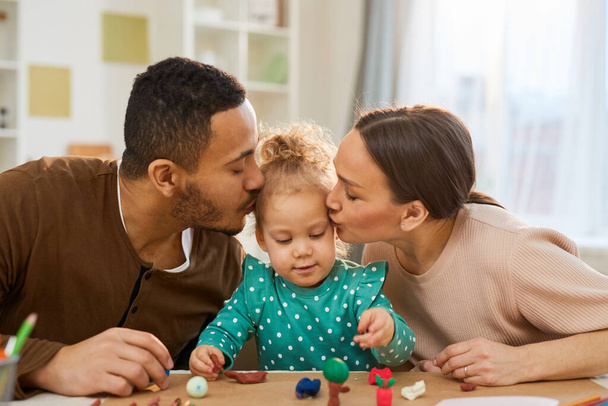 Young father and mother kissing their cute little daughter while she is making play dough shapes, horizontal shot - Foto, Imagem