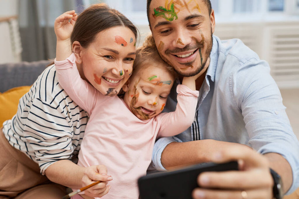 Jeune mari et femme prenant selfie drôle sur smartphone avec leur petite fille avec gouache sur le visage - Photo, image