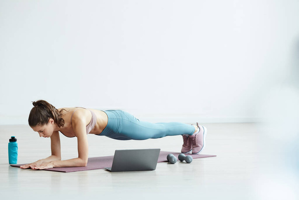 Minimal full length portrait of fit young woman doing plank exercise while watching workout video via laptop at home, copy space - 写真・画像