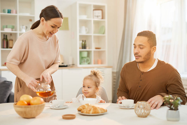 Junge Frau gießt beim Frühstück zu Hause Tee in Tassen für ihre kleine Tochter und ihren Mann - Foto, Bild