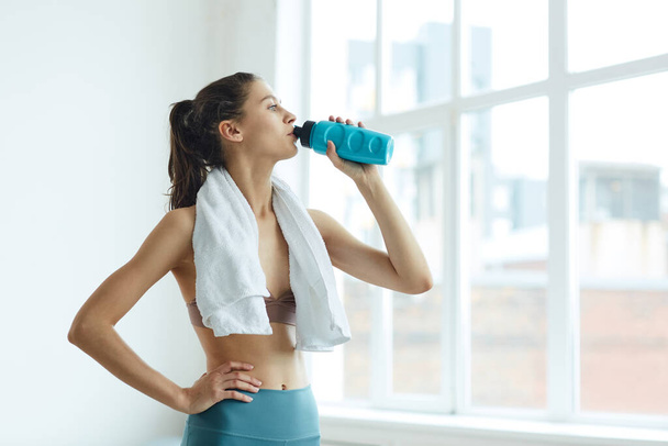 Cintura mínima para cima retrato de ajuste jovem mulher bebendo água enquanto em pé pela janela após o treino desportivo, espaço de cópia
 - Foto, Imagem