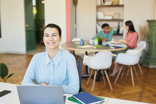 Весела молода бізнес-леді сидить за офісним столом з ноутбуком на ньому, посміхаючись на камеру, її колеги працюють за нею
 - Фото, зображення