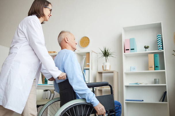 Visão lateral retrato de sorrir médico empurrando homem idoso em cadeira de rodas para exame ou consulta na clínica, espaço de cópia
 - Foto, Imagem