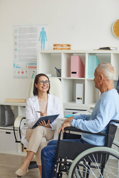 Portret van een lachende vrouwelijke arts die een gehandicapte oudere man in een rolstoel adviseert tijdens een consult in de kliniek - Foto, afbeelding