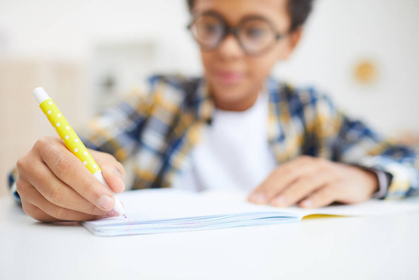 Nahaufnahme eines süßen afrikanischen Jungen, der während der Hausaufgaben in sein Notizbuch schreibt, sich auf den Vordergrund konzentriert, Raum kopiert - Foto, Bild