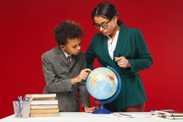 Portrait of young female teacher and cute African-American boy looking at geographical globe while standing against red background, copy space - Zdjęcie, obraz