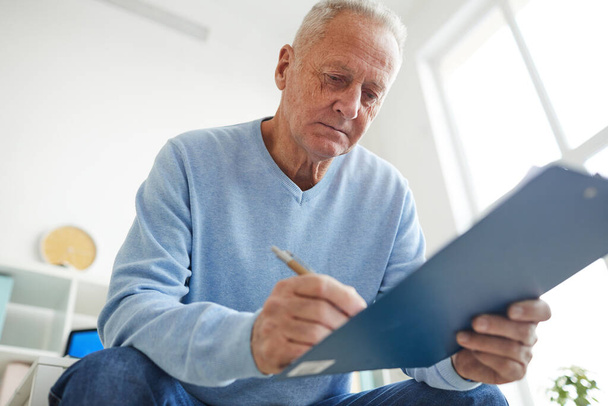 Nízký úhel pohledu na staršího muže držícího schránku a vyplňující formulář pacientů při registraci ke konzultaci na klinice - Fotografie, Obrázek