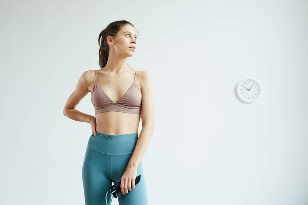 Minimal waist up portrait of fit young woman holding skipping rope and looking away while enjoying fitness workout standing against white wall with analog clock, copy space - Photo, Image
