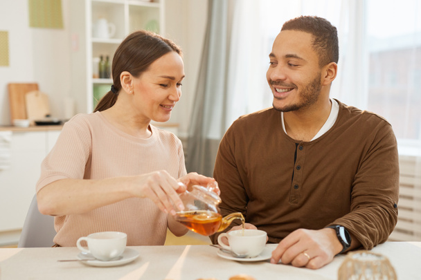 Horizontale mittlere Aufnahme einer verliebten jungen Frau, die ihrem glücklichen Mann während des Frühstücks Tee schenkt - Foto, Bild
