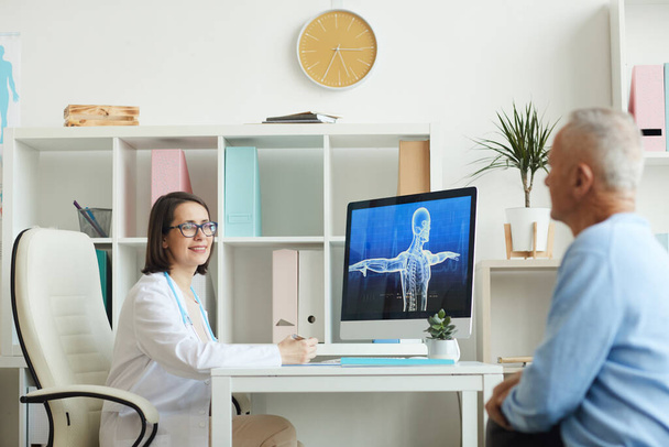 Ritratto di medico donna sorridente che consulta il paziente anziano mentre si siede alla scrivania in un moderno interno della clinica, copiare lo spazio - Foto, immagini