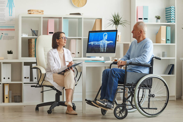 Retrato completo de homem idoso deficiente em cadeira de rodas conversando com médico feminino durante consulta na clínica
 - Foto, Imagem