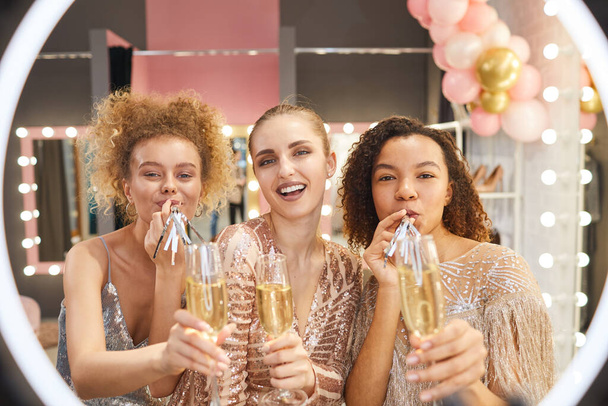 Grupo multi-étnico de mulheres jovens glamourosas segurando copos de champanhe e soprando chifres de festa enquanto olha para a câmera no interior do vestiário, filmado através da luz do anel
 - Foto, Imagem