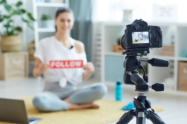Blurred portrait of smiling young woman holding FOLLOW sign while recording workout video at home and looking at camera, focus on foreground, copy space - Photo, Image