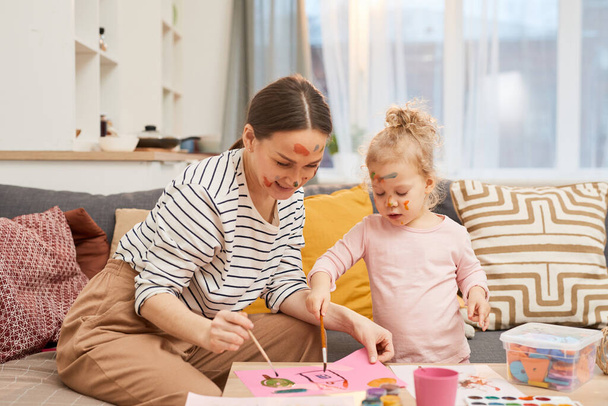 Jovem mãe alegre e sua filhinha gastando tempo livre pintando imagens coloridas usando guache
 - Foto, Imagem