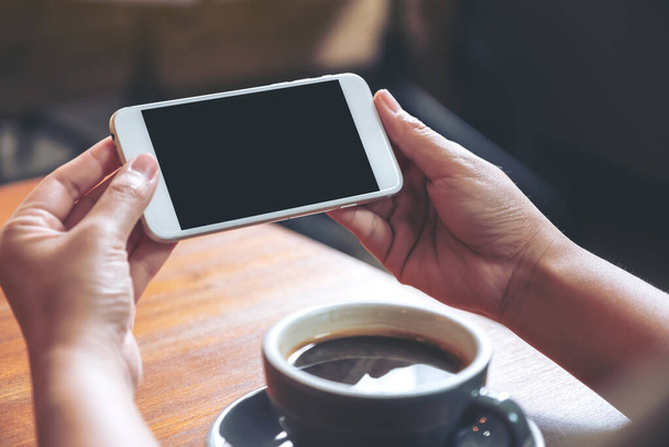 Образец изображения рук, держащих и использующих белый мобильный телефон с пустым черным экраном горизонтально для просмотра с чашкой кофе на деревянном столе
 - Фото, изображение