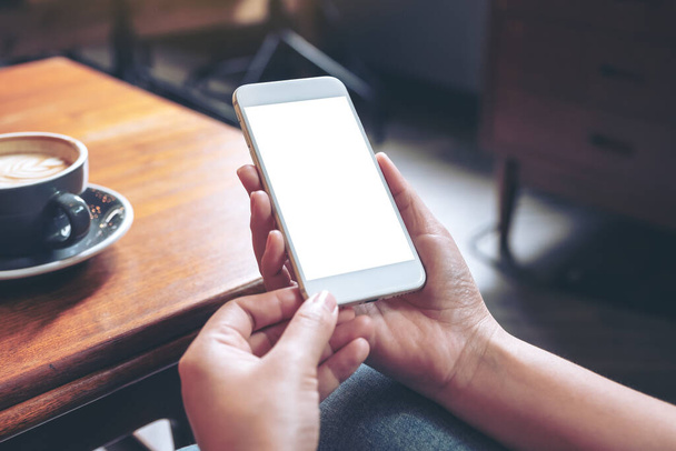 Image de maquette d'une main tenant un téléphone portable blanc avec écran de bureau vierge avec tasse à café sur une table en bois dans un café - Photo, image