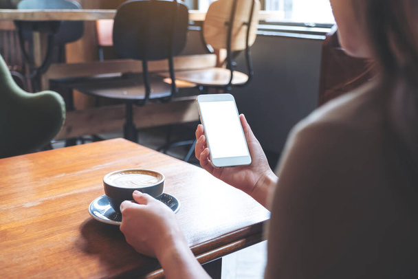 Mockup obraz ženy držící bílý mobilní telefon s prázdnou obrazovkou při pití kávy v moderní kavárně - Fotografie, Obrázek