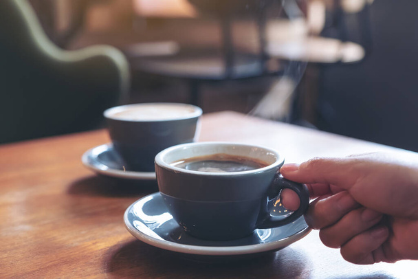 Closeup image of a hand holding a blue cup of hot coffee with smoke on wooden table in cafe - Photo, Image