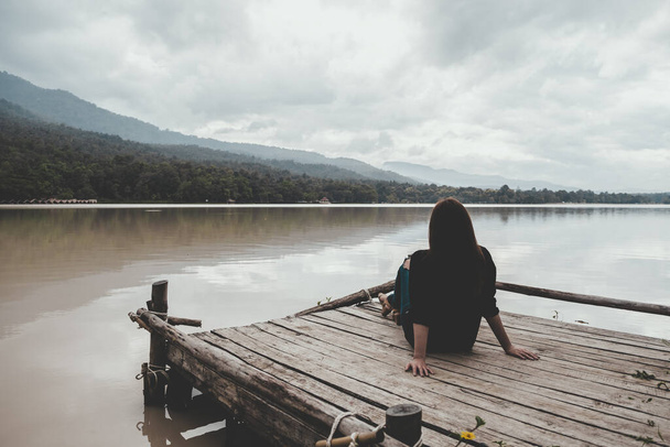 Closeup image of an asian woman sitting alone on an old wooden pier by the river with sky and mountain background - Photo, Image