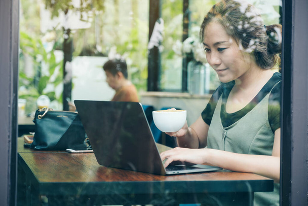 Asyalı bir iş kadınının kafeteryada kahve içerken bilgisayarını ahşap masada çalışırken ve kullanırken yakın plan görüntüsü. - Fotoğraf, Görsel
