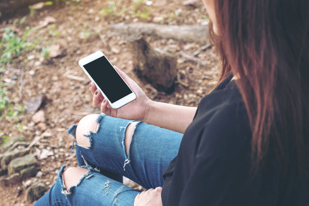 Imagen burlona de la mujer sosteniendo el teléfono móvil blanco con pantalla de escritorio negro en blanco mientras está sentado al aire libre
 - Foto, Imagen