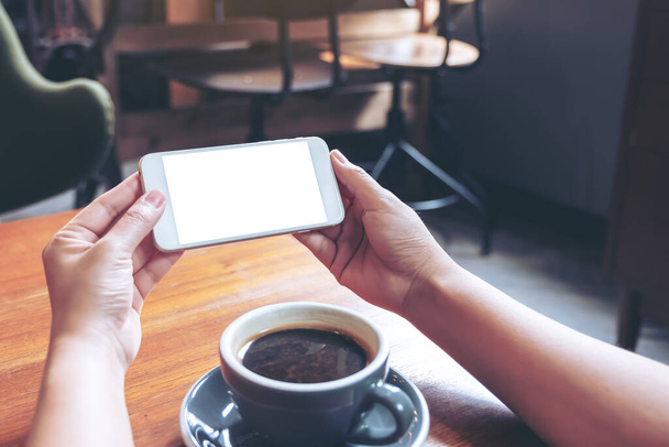 Образец изображения рук, держащих и использующих белый мобильный телефон с пустым экраном горизонтально для просмотра с чашкой кофе на деревянном столе
 - Фото, изображение
