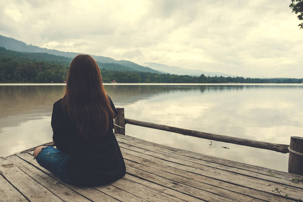 Imagen de cerca de una mujer asiática sentada sola en un viejo muelle de madera junto al río con el cielo y el fondo de la montaña
 - Foto, Imagen