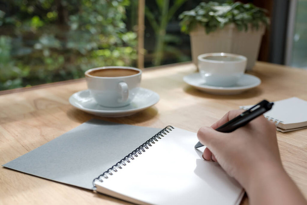 Nahaufnahme einer Frau, die auf ein weißes Blanko-Notizbuch mit Kaffeetasse auf einem Holztisch schreibt  - Foto, Bild