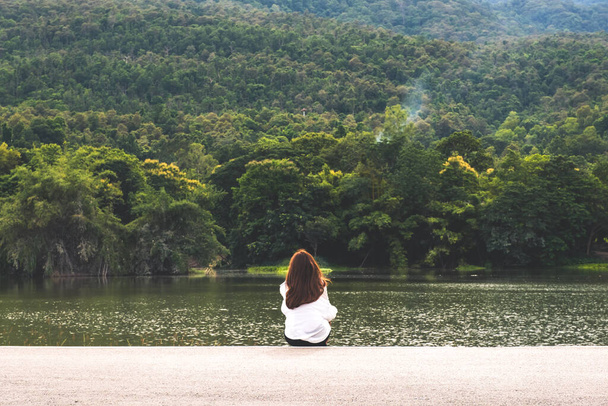 Μια γυναίκα κάθεται μόνη της στη λίμνη κοιτάζοντας τα βουνά με συννεφιά και πράσινο φόντο της φύσης - Φωτογραφία, εικόνα