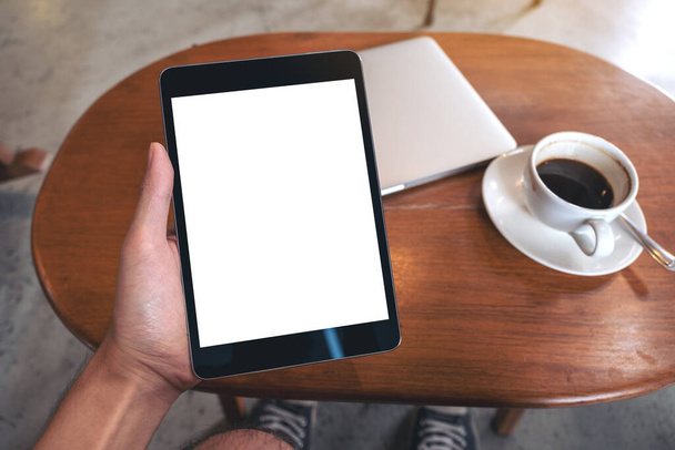 Mockup afbeelding van de hand houden zwarte tablet pc met blanco desktop wit scherm met laptop en koffiekopje op houten tafel - Foto, afbeelding