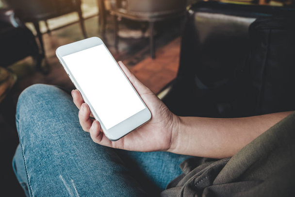 Mockup-Bild der Hand einer Frau, die ein weißes Mobiltelefon mit leerem Desktop-Bildschirm am Oberschenkel hält, während sie im Café sitzt - Foto, Bild