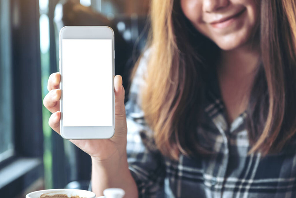 Attrappe einer Frau, die ein weißes Handy mit leerem Bildschirm in einem modernen Café hält und zeigt - Foto, Bild