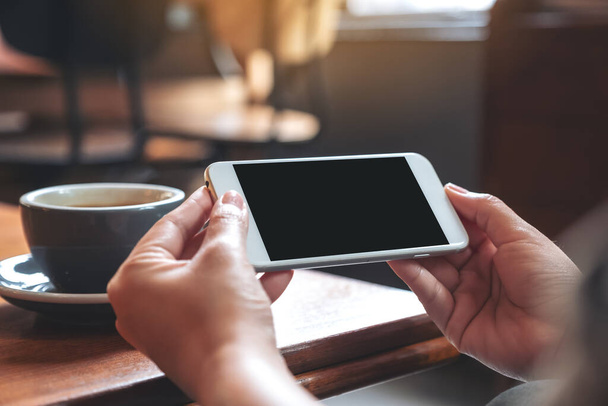 Образец изображения рук, держащих и использующих белый мобильный телефон с пустым черным экраном горизонтально для просмотра с чашкой кофе на деревянном столе
 - Фото, изображение