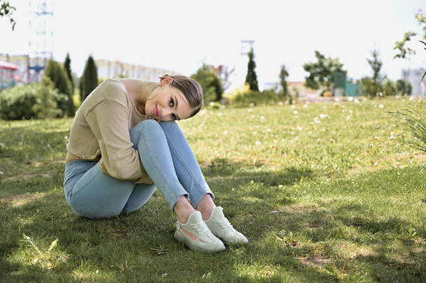 Una modelo se sienta en el suelo en un parque de verano con un clima soleado. Retrato de una chica caucásica con maquillaje de belleza
 - Foto, Imagen