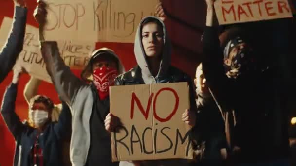 Mulher branca bonita no capuz em pé na multidão de pessoas em protesto de rua com cartaz Nenhum racismo à noite. Manifestação dos EUA por direitos humanos iguais. Protestante feminina
. - Filmagem, Vídeo