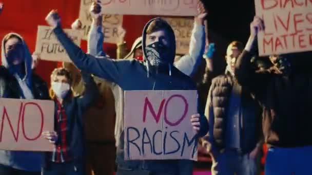 Jovem caucasiano de capuz em pé na multidão de pessoas em protesto de rua com cartaz Sem racismo à noite. Manifestação dos EUA por direitos humanos iguais. Protestante masculino. Ativista e líder
. - Filmagem, Vídeo