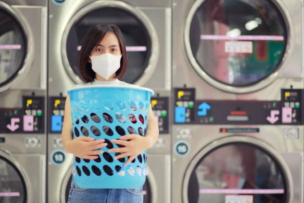 Corona virüsü COVID-19 salgını için karantinada maske takan bir kadın. Çamaşırhanede çamaşır yıkarken. Enfeksiyon riski için yeni normal yaşam tarzı sosyal mesafesi. - Fotoğraf, Görsel