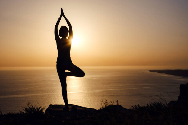 A nő jóga testtartással a hegyen naplementekor Silhouette gyönyörű fiatal nő gyakorló jóga szabadban. wellness, egészségügyi, sport és életmód koncepció - Fotó, kép