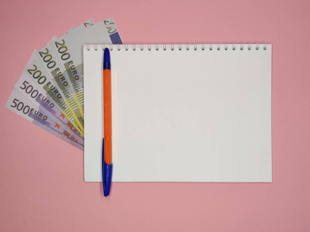 on a pink background, a pen on a Notepad with a copy space, 200 and 500 Euro bills peek out from under it. horizontal photo. - Foto, afbeelding