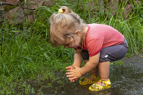 A little girl stands with her feet in a puddle and plays with water, CHILDLIKE SPONTANEITY, - Foto, afbeelding