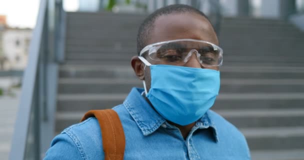 Close up of happy face of young handsome African American man in medical mask and goggles outdoors. Portrait shot of cheerful male wearing respiratory protection at street. - Imágenes, Vídeo