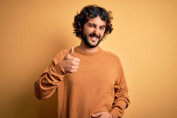 Ein junger gutaussehender Mann mit Bart und lässigem Pullover steht vor gelbem Hintergrund und macht eine glückliche Daumen-nach-oben-Geste mit der Hand. Zustimmender Blick in die Kamera zeigt Erfolg. - Foto, Bild