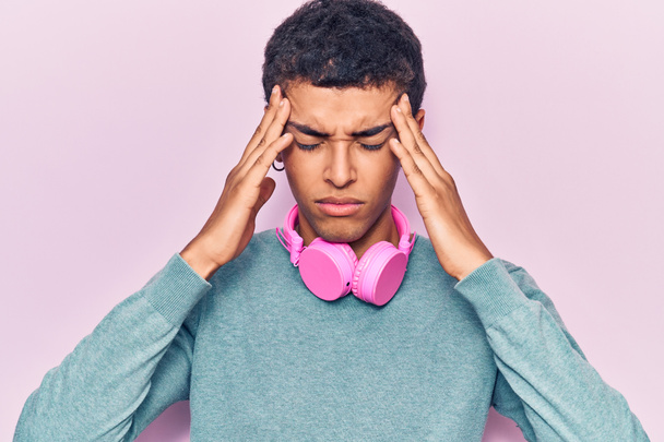 Jonge Afrikaans-Amerikaanse man luistert naar muziek met behulp van koptelefoons die lijden aan hoofdpijn wanhopig en gestresst omdat pijn en migraine. handen op het hoofd.  - Foto, afbeelding