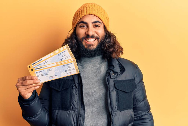 Young arab man holding boarding pass looking positive and happy standing and smiling with a confident smile showing teeth  - Photo, Image