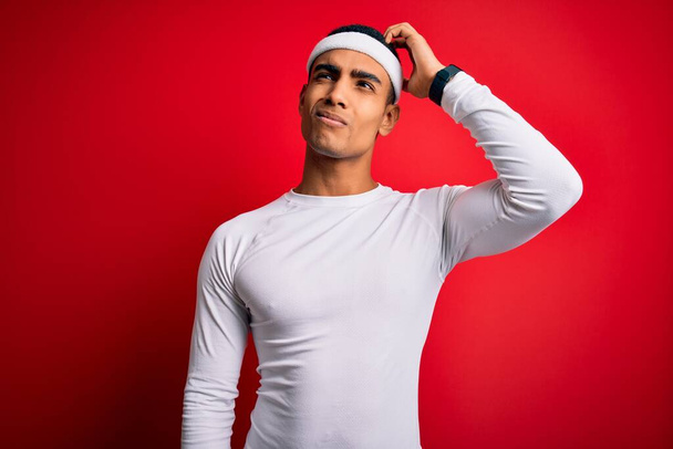 Young handsome african american sportsman wearing sportswear over red background confuse and wondering about question. Uncertain with doubt, thinking with hand on head. Pensive concept. - Photo, Image