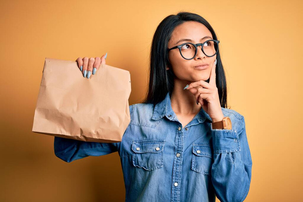 Giovane bella donna cinese che tiene borsa di consegna su sfondo giallo isolato faccia seria pensando alla domanda, idea molto confusa
 - Foto, immagini